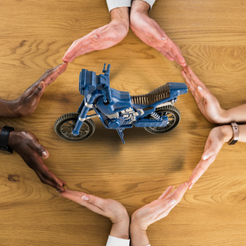Alt text: A top-down view of a table where four people have joined hands in a circle around a toy motorcycle, symbolizing the comprehensive protection offered by motorcycle insurance.